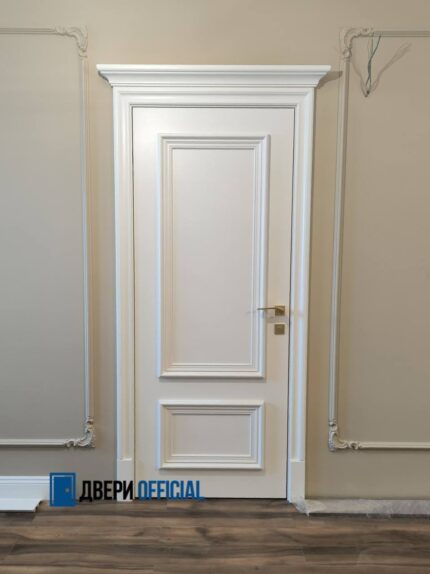 Белая дверь с двумя прямоугольными декоративными рамками, широкими наличниками и верхним карнизом. На фотографии установлена ручка золотистого оттенка.